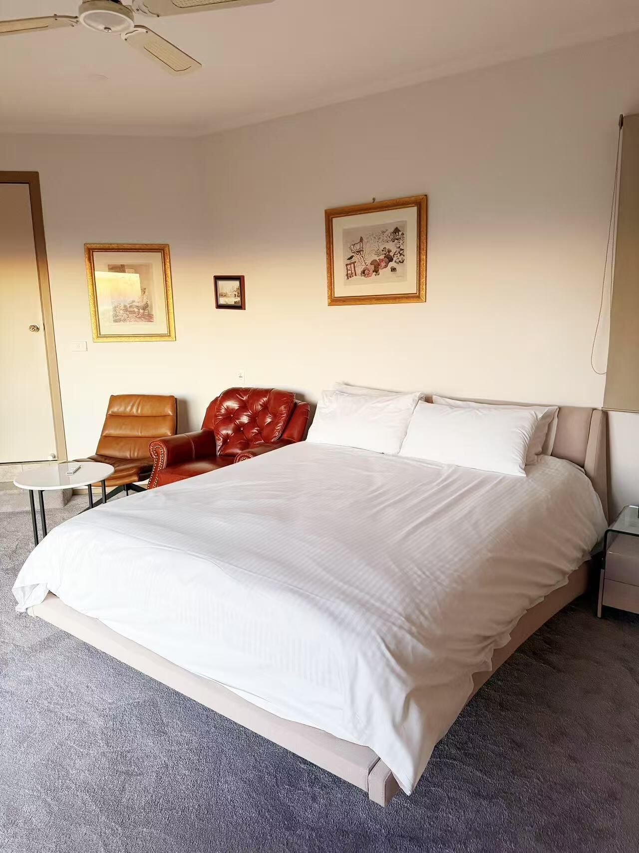 A hotel room with a large bed, a red leather chair, a small round table, and two framed pictures on the wall.