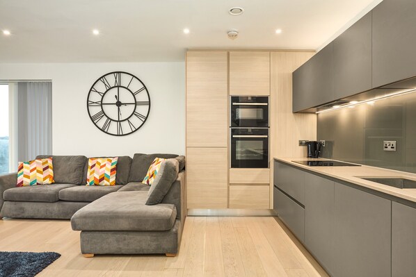 Eine moderne Küche mit integrierten Geräten, ein graues Sofa mit bunten Kissen und eine große Wanduhr.