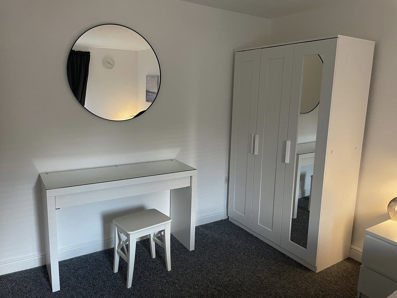 Une pièce avec un bureau blanc, un petit tabouret, une armoire et un miroir rond accroché au mur.
