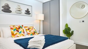 Een net opgemaakt bed met een blauw-witte deken, een kleurrijk kussen en een stapel gevouwen witte handdoeken. Boven het bed hangt een klok en een kunstwerk in een lijst.