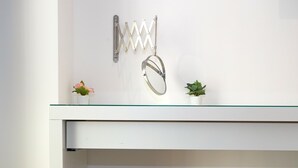 Een wit bureau met een glazen blad, een klein wit bankje en een metalen rek met een spiegel en twee potplanten.