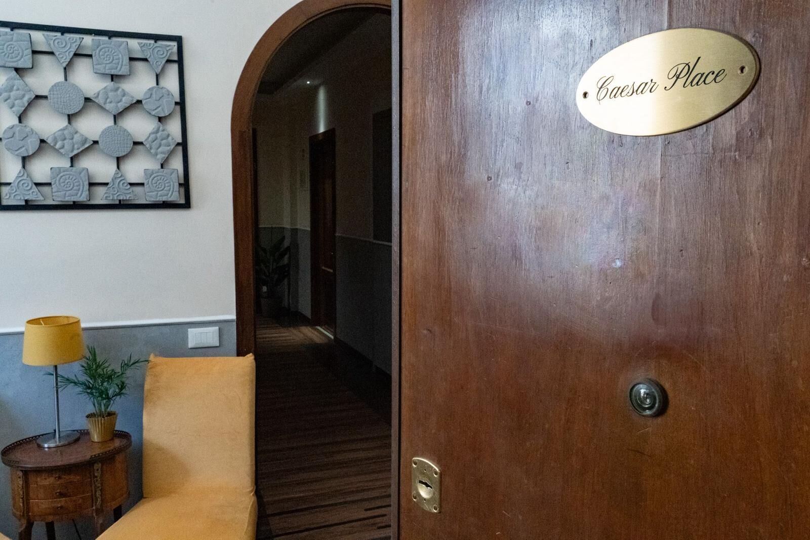 A hotel lobby with a wooden door labeled "Caesar Place," a small table with a lamp, and a decorative wall piece.