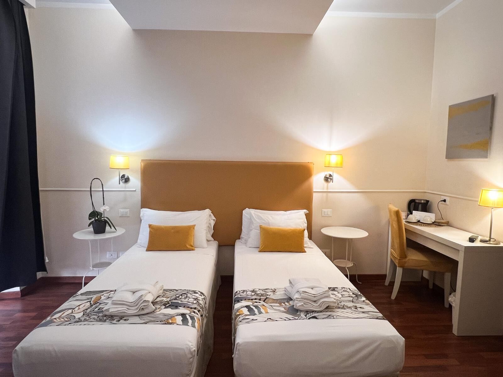 A hotel room with two beds, a desk, a chair, a small table, and a potted plant.