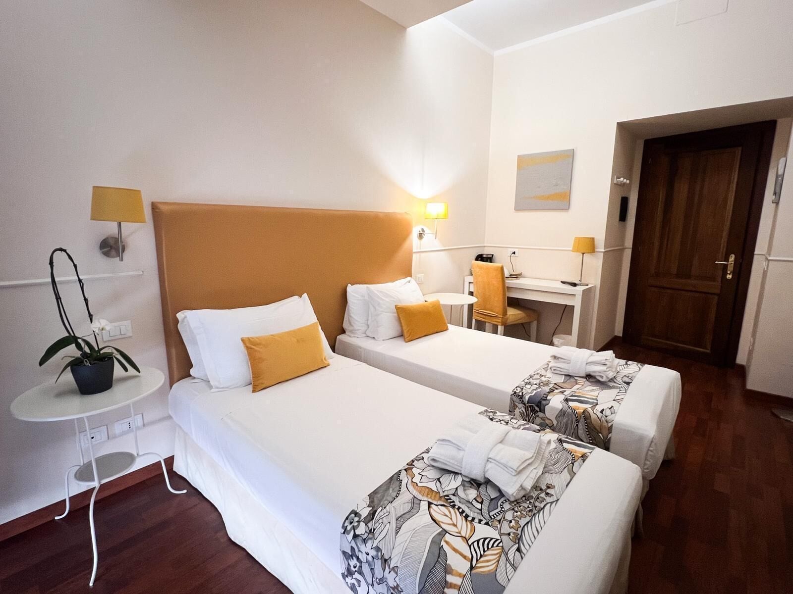 Een hotelkamer met twee bedden, een bureau, een stoel, een klein tafeltje met een lamp en een potplant.