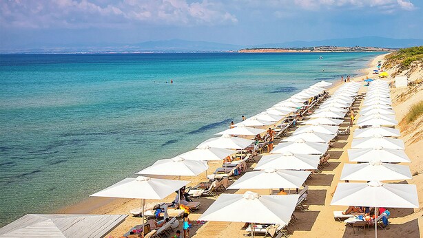 A beach with rows of white umbrellas and sun loungers, a clear blue sea, and a distant shoreline with buildings.