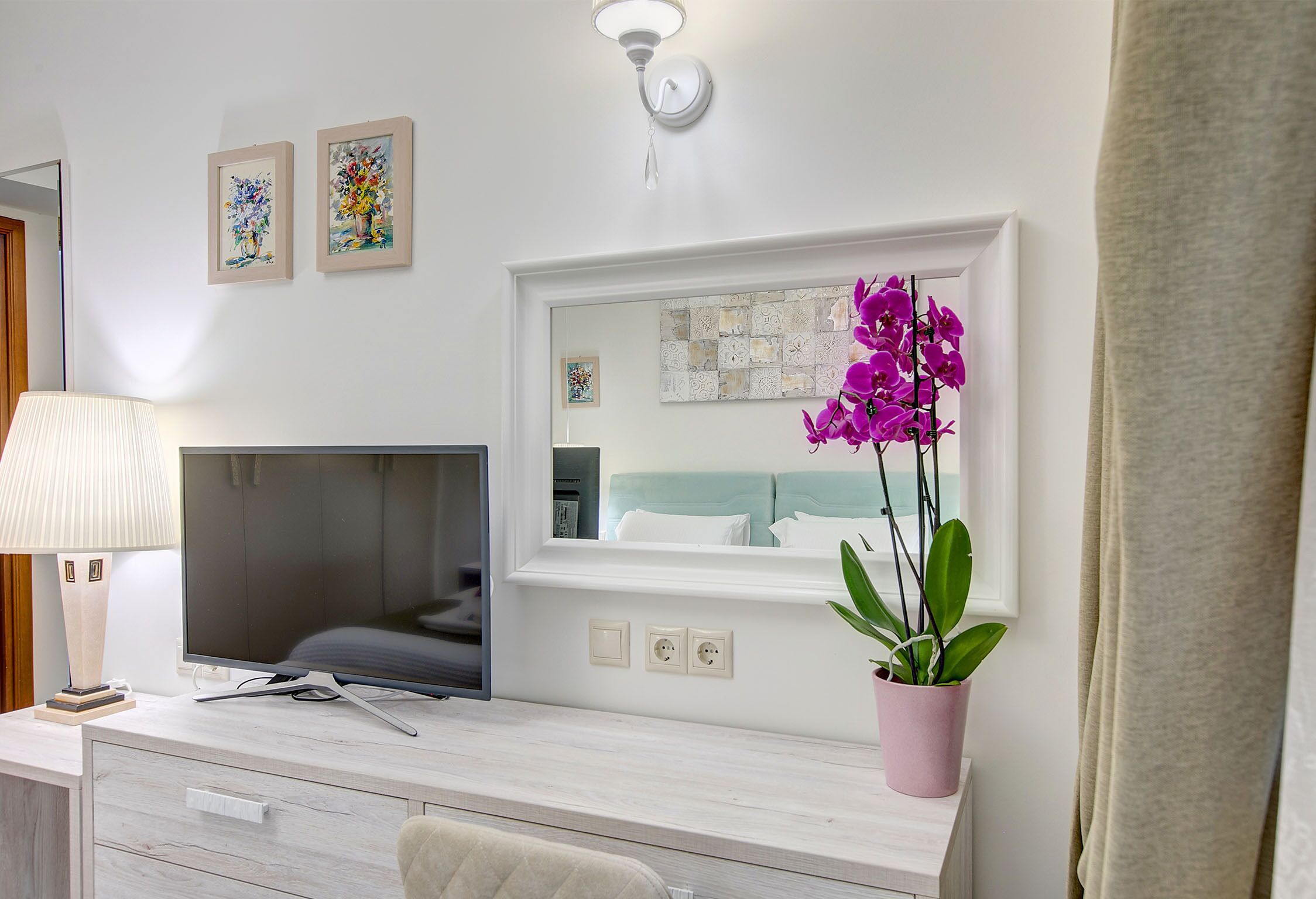 A hotel room with a TV, a desk with a lamp, a mirror, and a potted orchid.