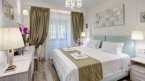 A hotel room with a large bed, two bedside tables, a desk, a chair, a desk lamp, a wall-mounted lamp, a chandelier, a window with curtains, and a decorative wall piece.