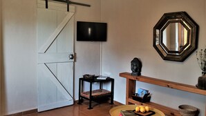 Ein Wohnzimmer mit Fernseher, einem runden Tisch mit Büchern und Obst, einer Couch, einem Holzregal mit Dekoration und einem großen Spiegel.