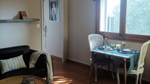 Ein Wohnzimmer mit einer Couch, einem Esstisch für zwei Personen, einer Hängelampe und einem Fenster mit Vorhängen.