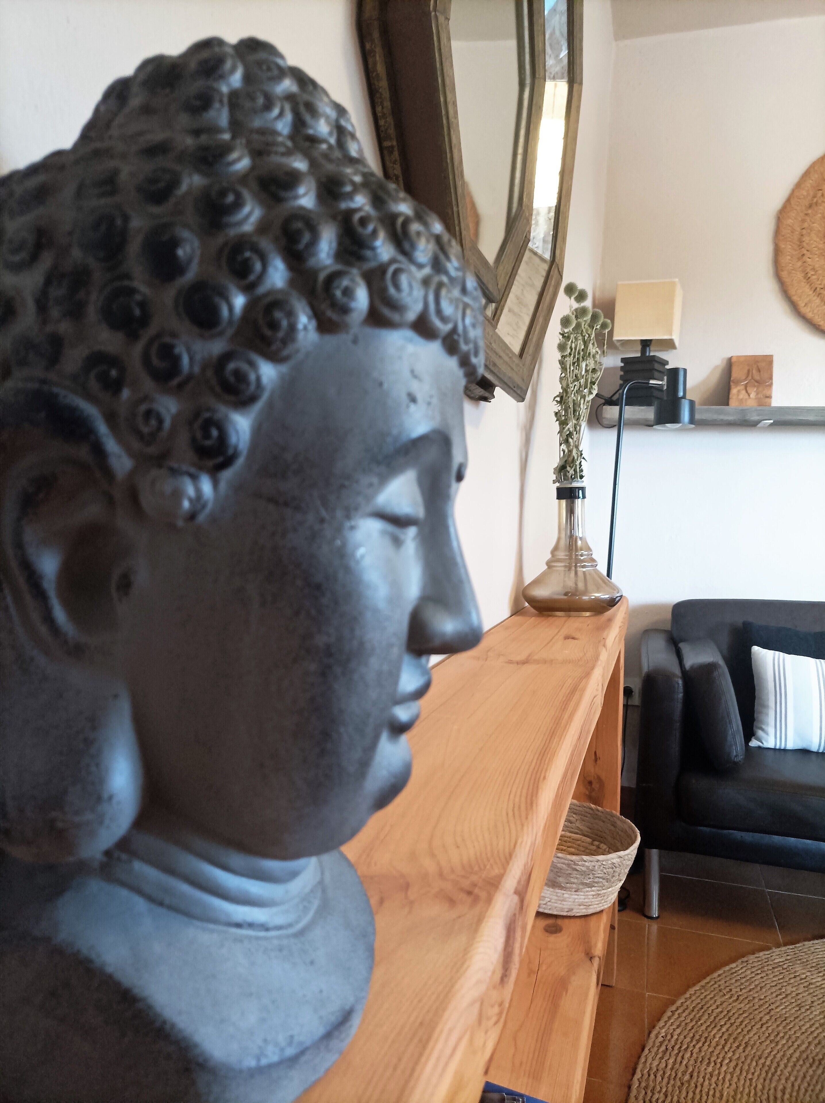 Eine Buddha-Statue auf einem Holzboden, ein Spiegel, eine Vase mit Trockenblumen, eine Lampe und ein dunkles Sofa mit Kissen.