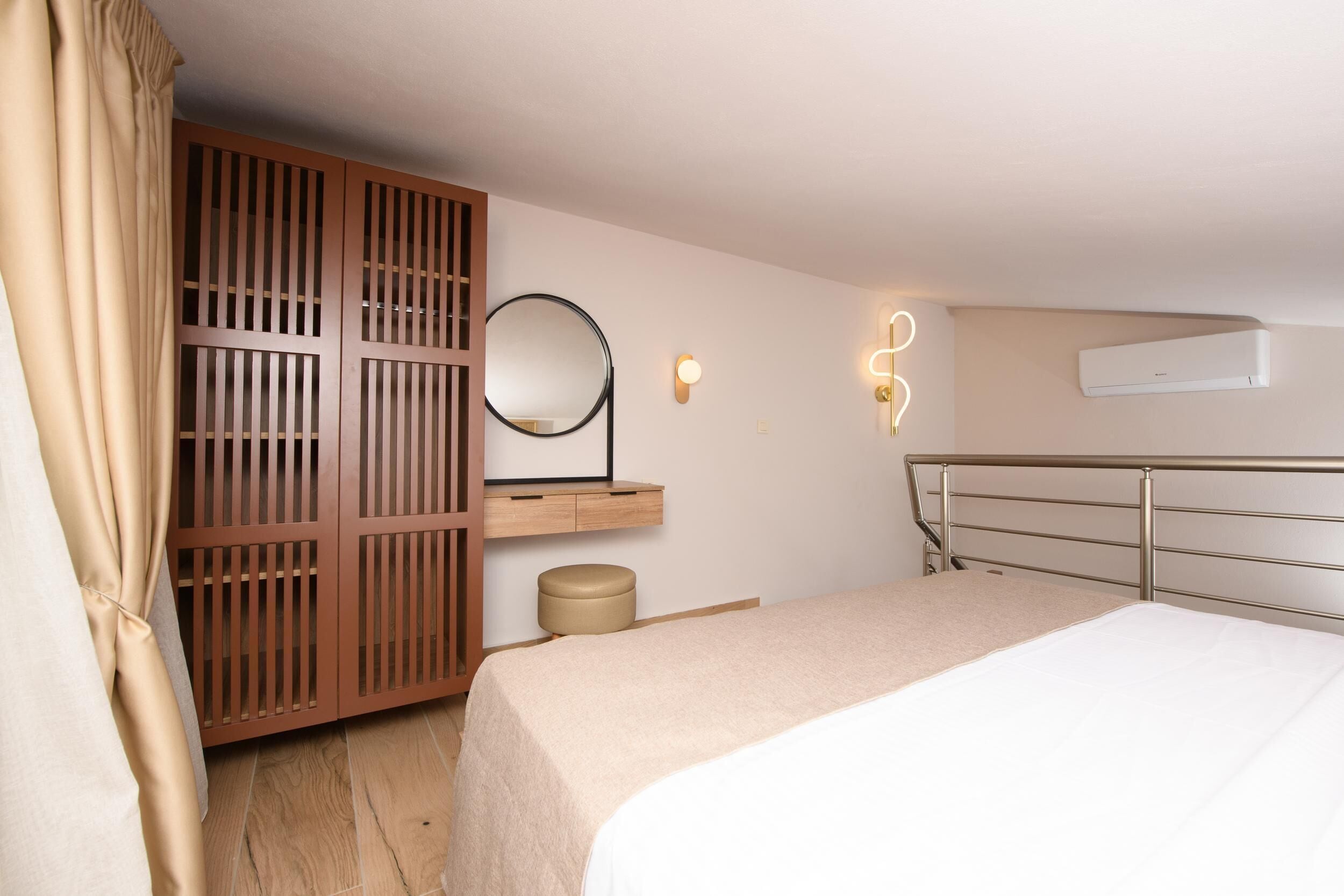 A bedroom with a bed, a wooden wardrobe, a round mirror, a bedside table, and an air conditioning unit.
