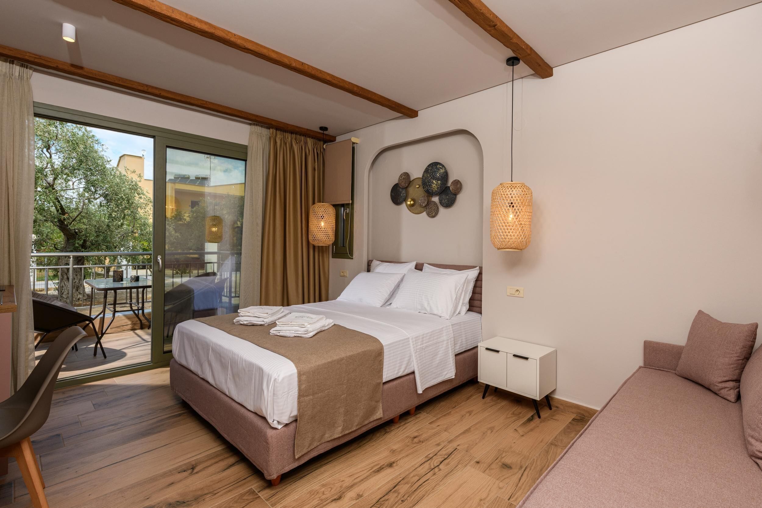 A modern bedroom with a large bed, a small bedside table, a sofa, a balcony with a table and chairs, and a view of trees and buildings outside.