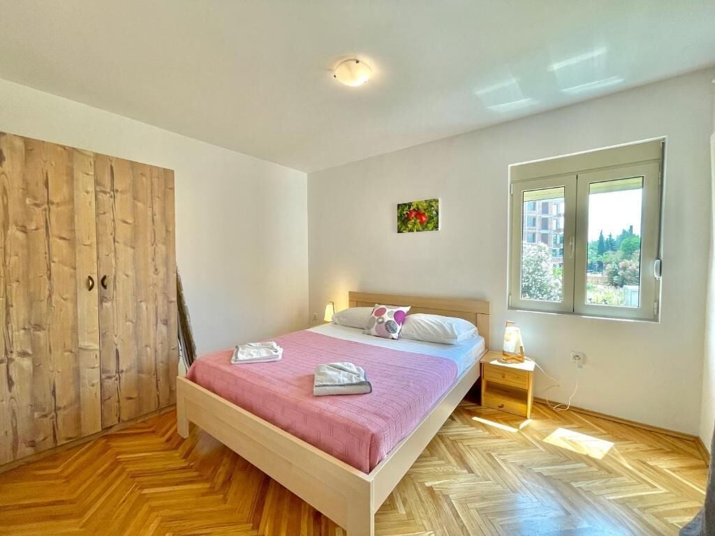 Een slaapkamer met een houten kledingkast, een bed met een roze dekbedovertrek, een nachtkastje met een lamp en een raam met uitzicht op gebouwen.