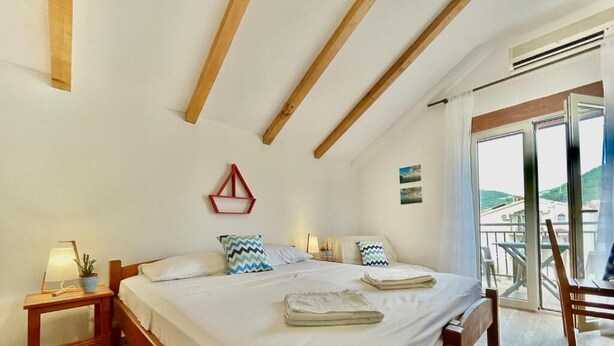 A bedroom with a wooden bed, a nightstand, a red geometric wall decoration, and a view of a balcony with outdoor furniture. - Garden House Budva (Budva)