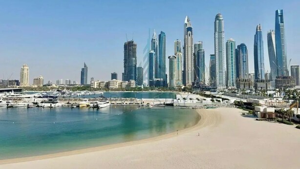 Un paisaje urbano costero con rascacielos modernos, un puerto deportivo y una playa de arena.
