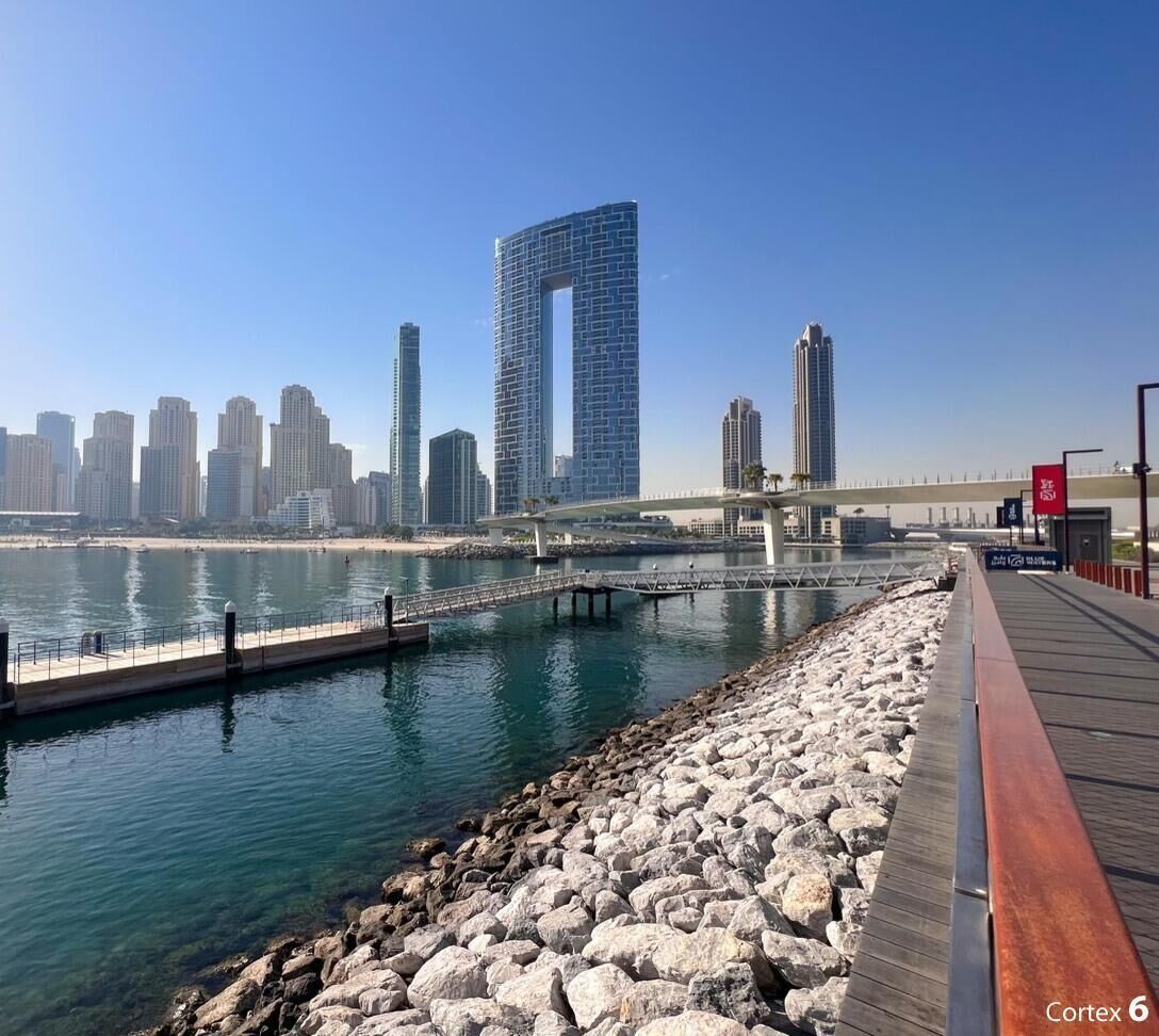 A waterfront promenade with a modern bridge, a marina, and a city skyline in the background.