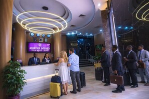 A hotel reception desk with a digital display, a waiting area, and a staircase leading to the upper floors.