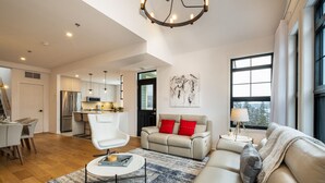 A spacious living room with a sofa, a coffee table, and a dining area with chairs. There is a kitchen in the background with modern appliances. - Tremblant Prestige - Les Eaux 215-8 (Mont-Tremblant)