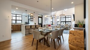 A spacious living area with a dining table set for four, a kitchen in the background, and large windows providing natural light. - Tremblant Prestige - Les Eaux 215-8 (Mont-Tremblant)