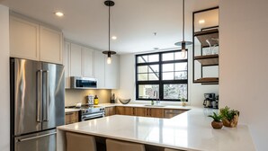 A modern kitchen with stainless steel appliances, a large island with bar stools, pendant lighting, and a large window providing natural light. - Tremblant Prestige - Les Eaux 215-8 (Mont-Tremblant)