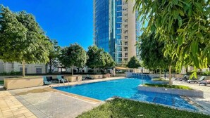 A swimming pool with lounge chairs, surrounded by greenery and a tall building in the background.
