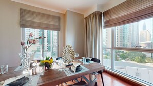 A dining area with a wooden table set for two, large windows with blinds, a view of high-rise buildings, and a decorative leaf centerpiece.