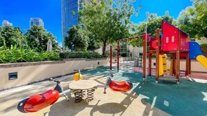 A playground with a swing set and climbing structures, surrounded by greenery and tall buildings.