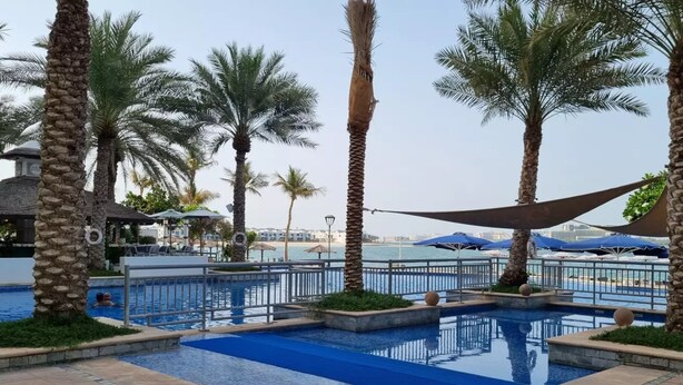 A pool area with palm trees, a clear sky, and a view of the sea. - Luxury 2BR | Palm Jumeirah | Pool Access (Dubai)