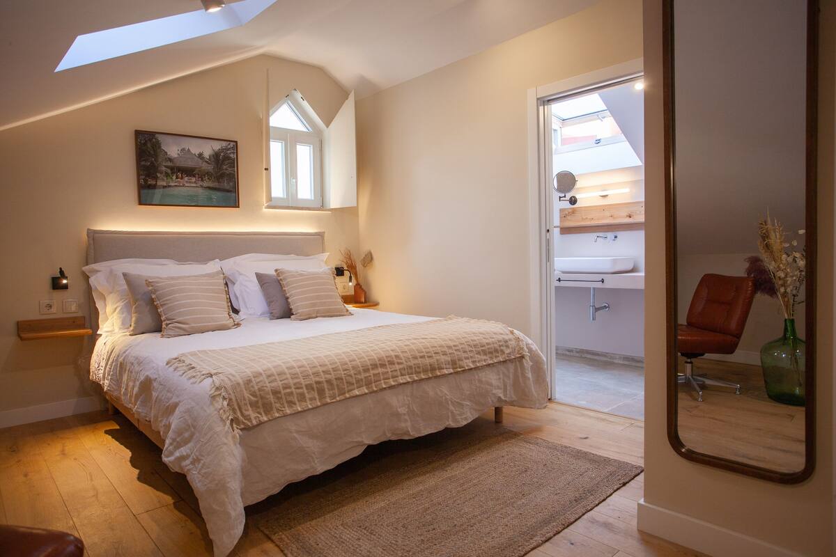 A bedroom with a large bed, a nightstand, a bedside table, a mirror, and a view of a bathroom.