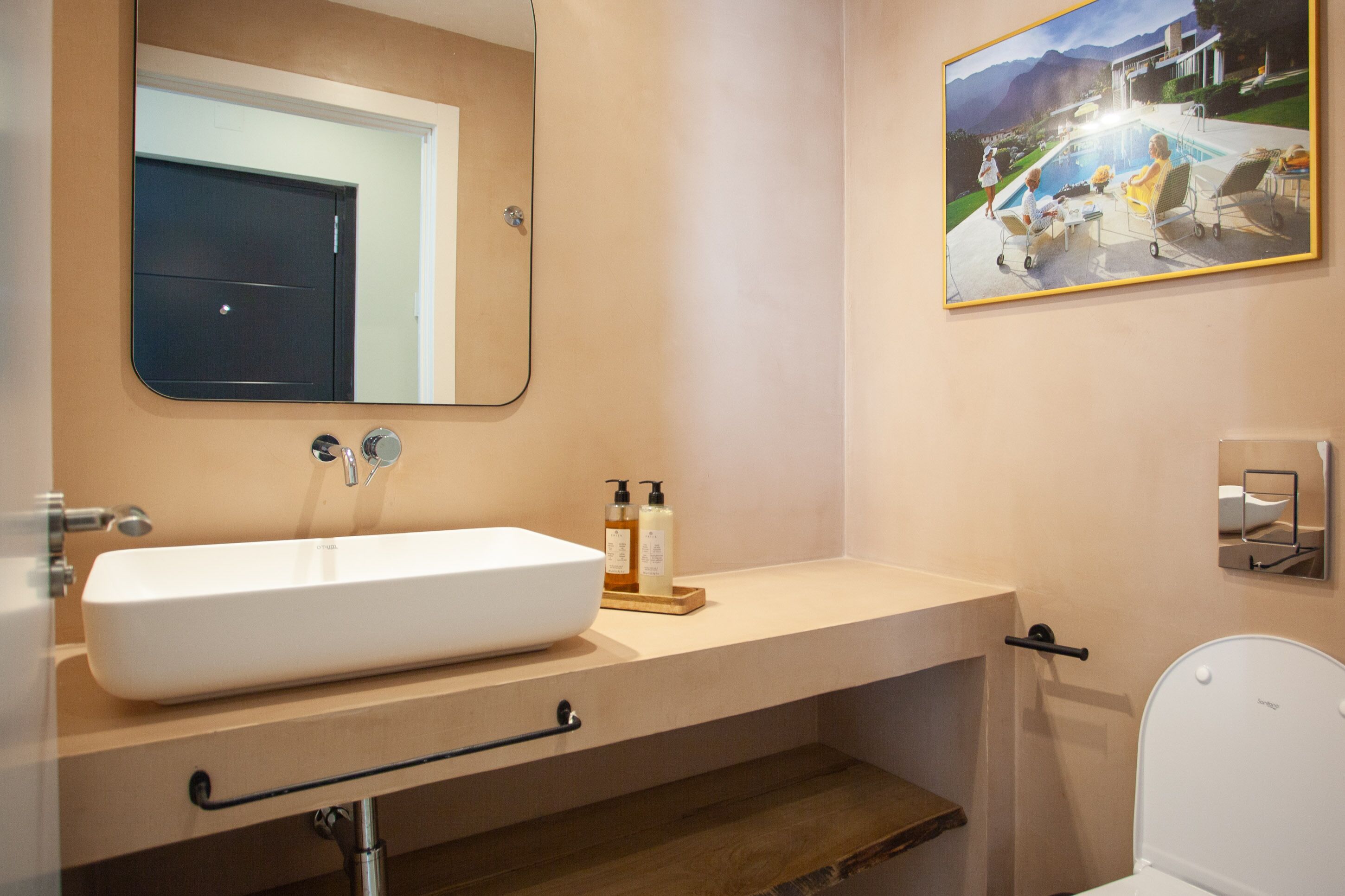 Une salle de bain avec un grand lavabo blanc, un miroir, un tableau représentant une famille au bord d’une piscine et des toilettes.