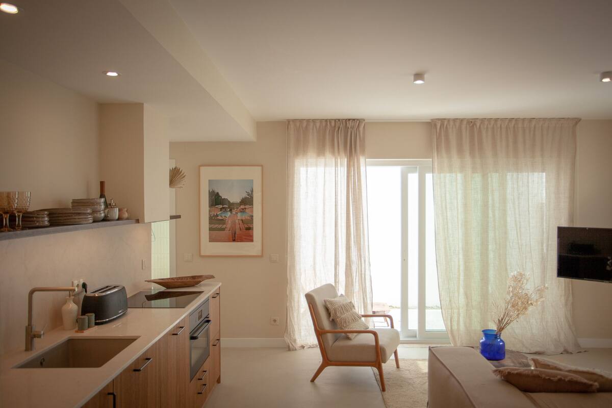 A modern living room with a kitchen area, a comfortable seating area, and a large window with curtains.