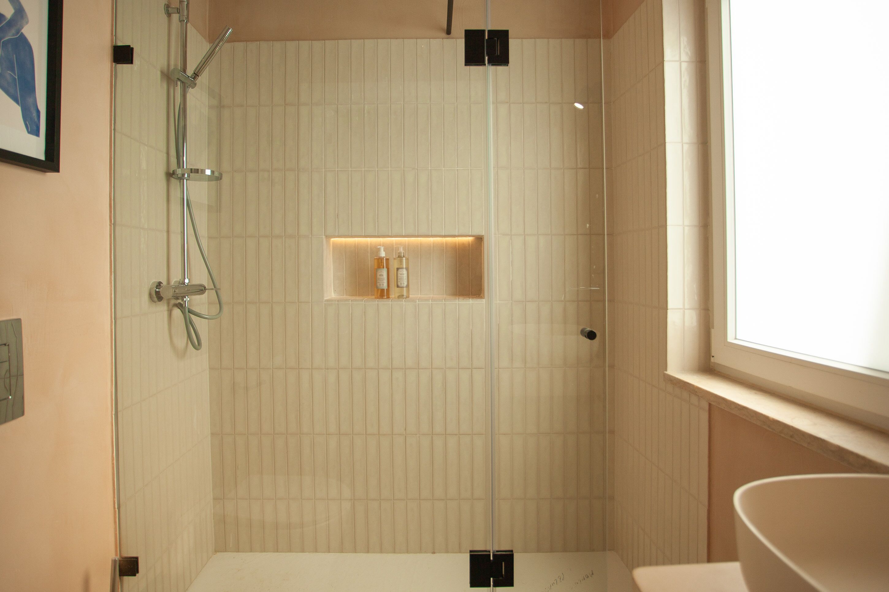 Un baño moderno con ducha envuelta en vidrio, repisa empotrada con artículos de aseo y un amplio ventanal que proporciona luz natural.