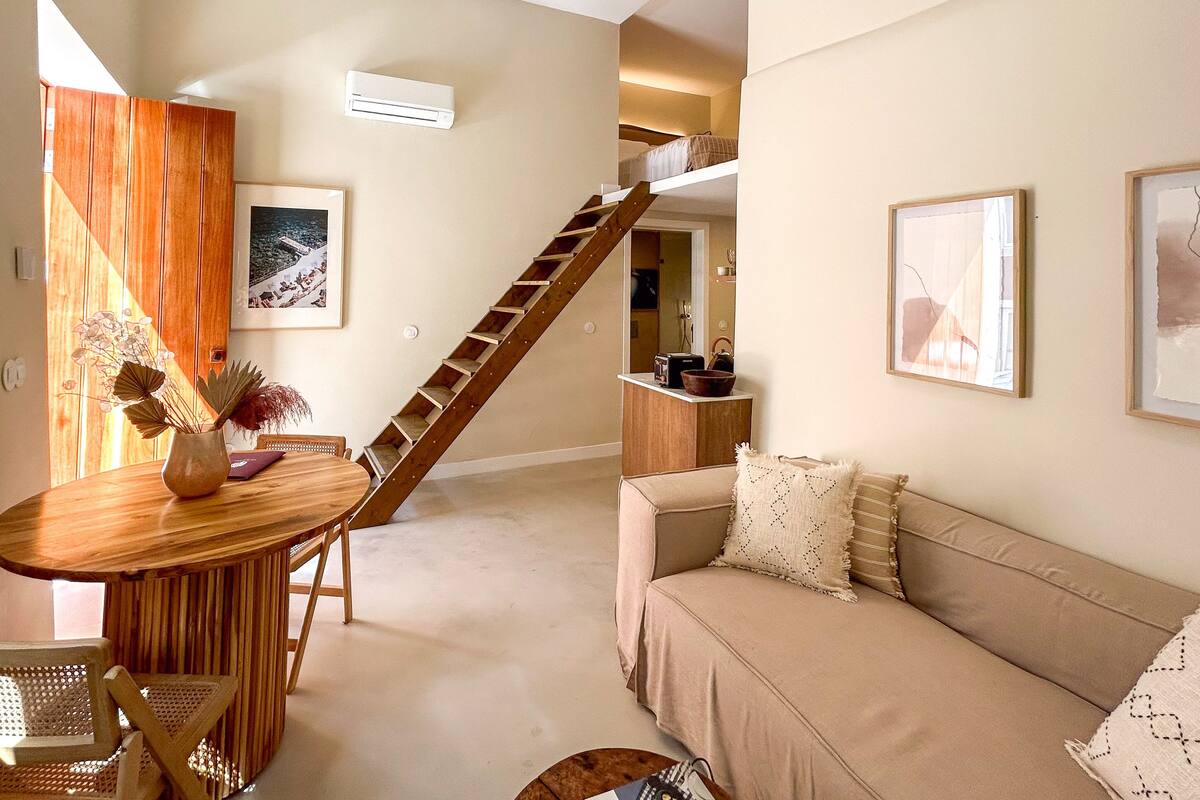 A modern living room with a wooden dining table, a sofa, a staircase, and a kitchen area.