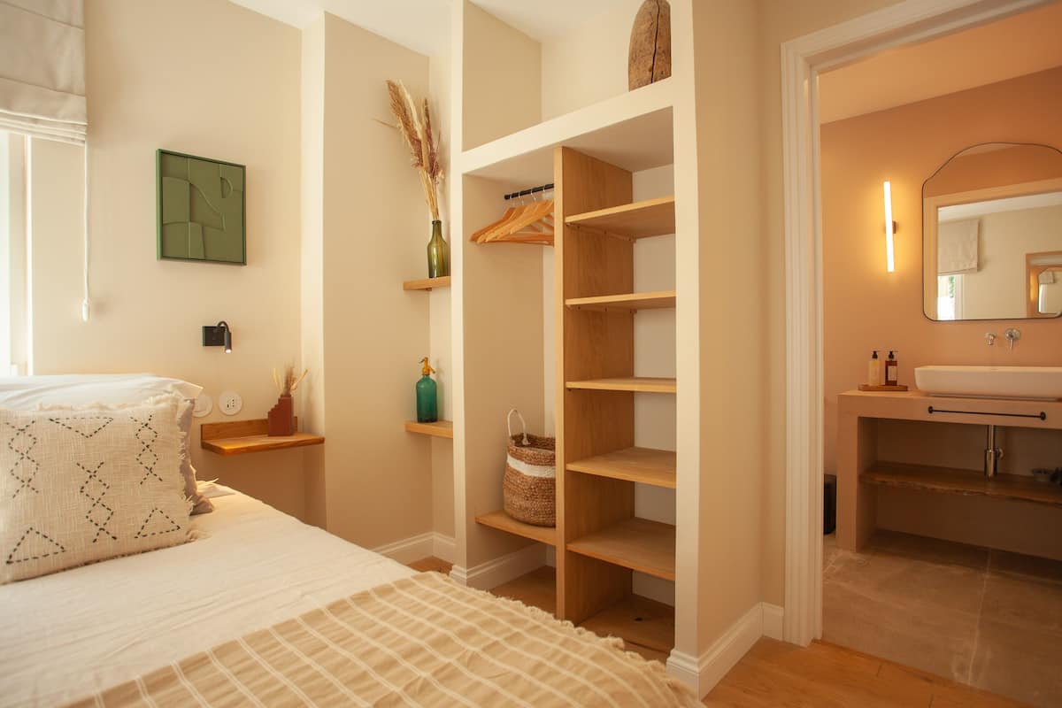A bedroom with a bed, a closet with shelves, a bedside table with a lamp, and a bathroom with a sink and mirror.