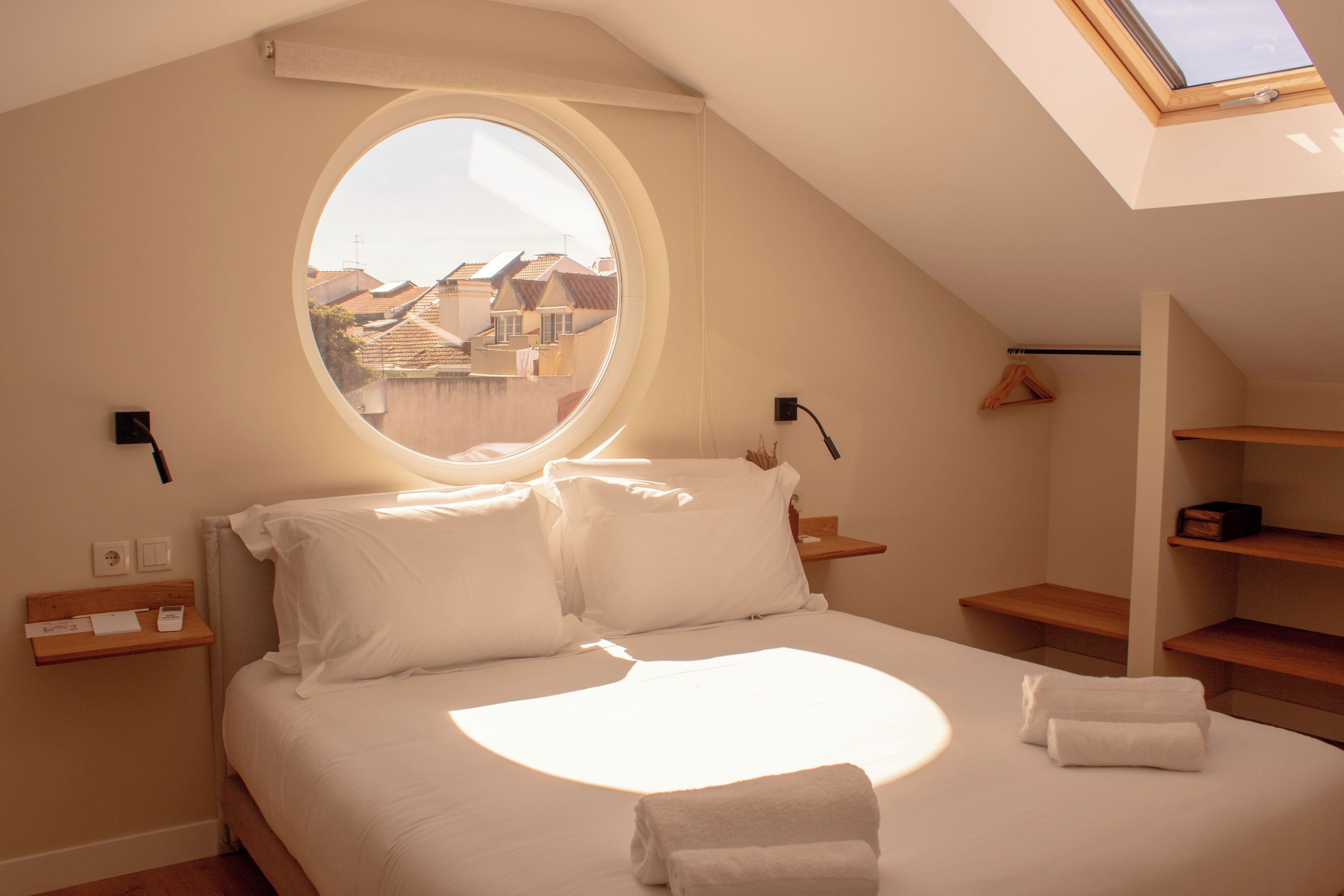 A bedroom with a skylight, a bed with pillows, a bedside table, and a view of buildings outside.