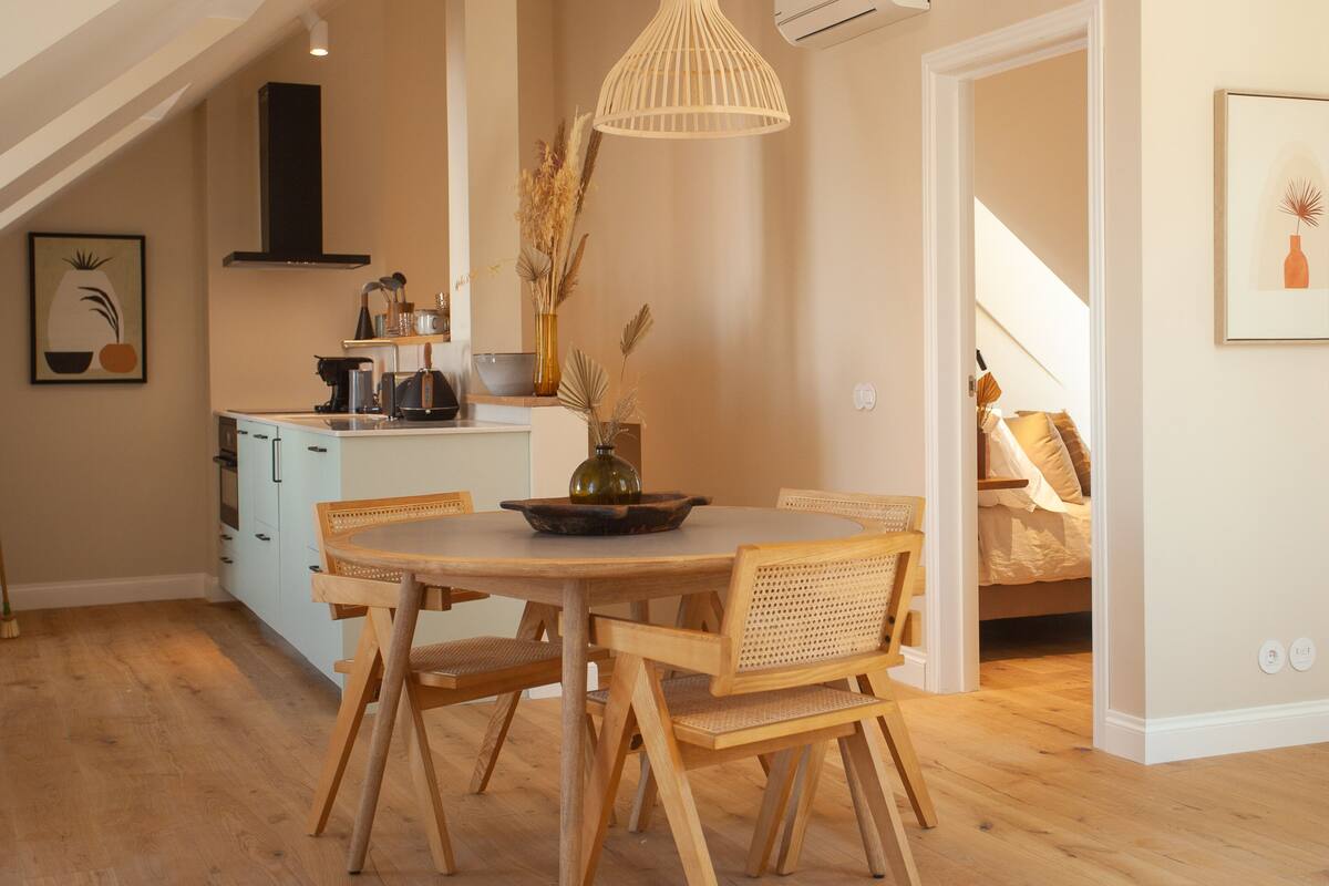 A dining area with a round wooden table and chairs, a hanging light fixture, and a kitchen in the background.