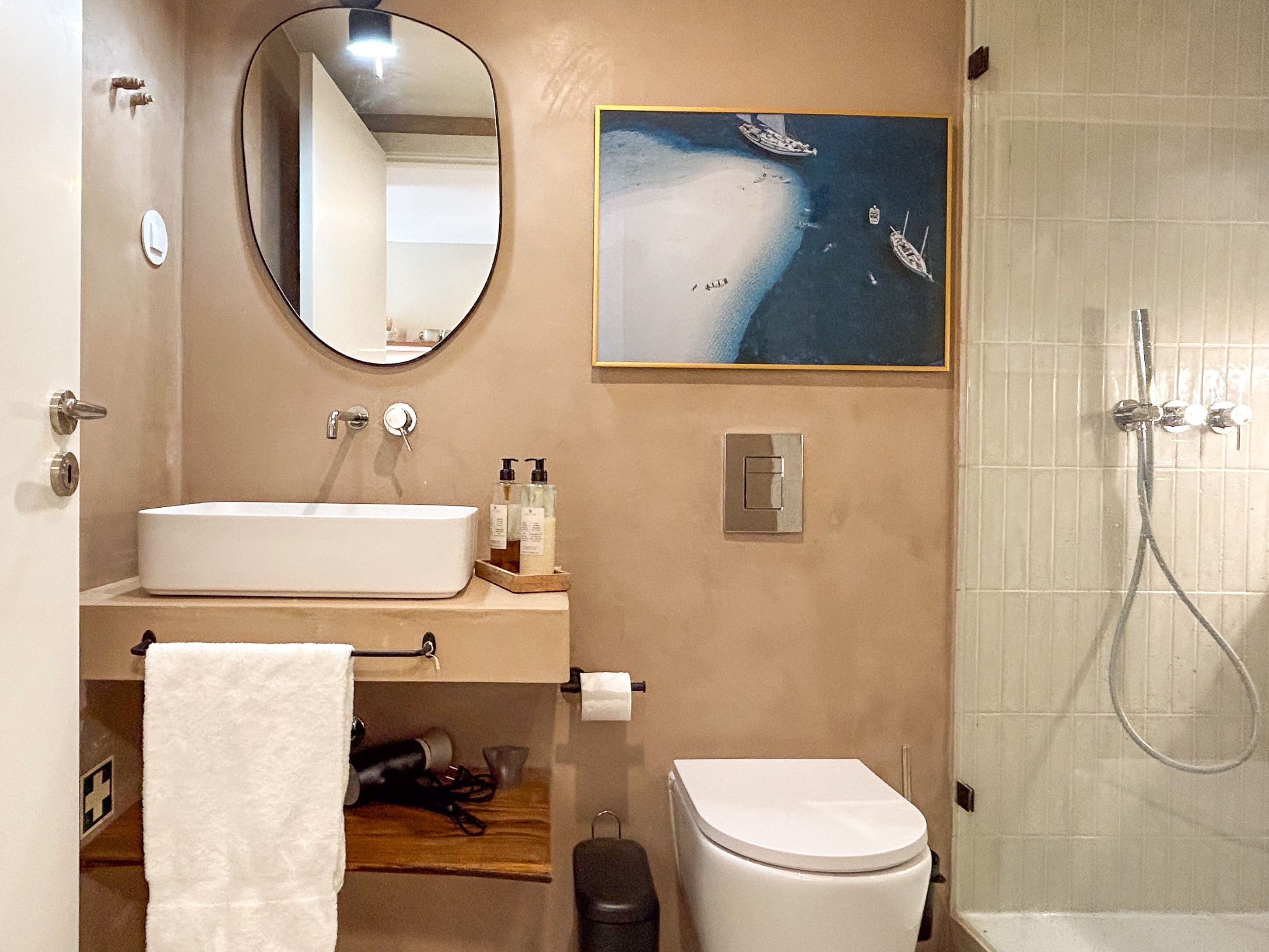 A bathroom with a white sink, mirror, toilet, and shower. There is a painting of boats on the wall.