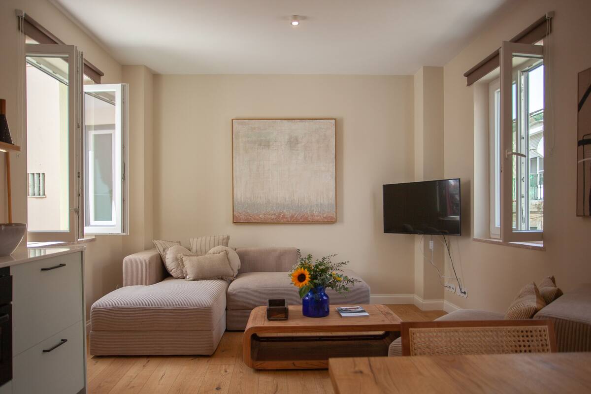 A modern living room with a sectional sofa, a coffee table with a vase of flowers, a flat-screen TV mounted on the wall, and large windows providing natural light.