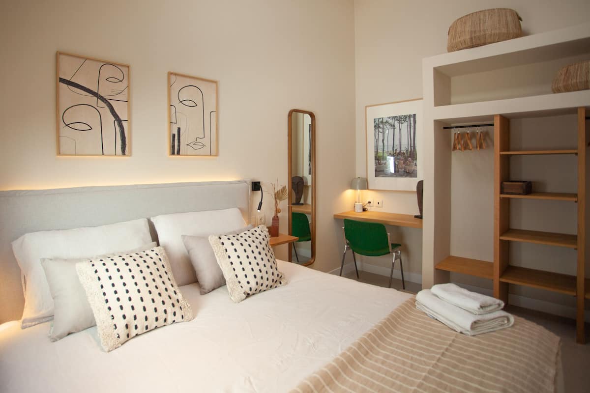 A neatly arranged hotel room with a bed, pillows, a desk, a chair, a mirror, and a wardrobe.