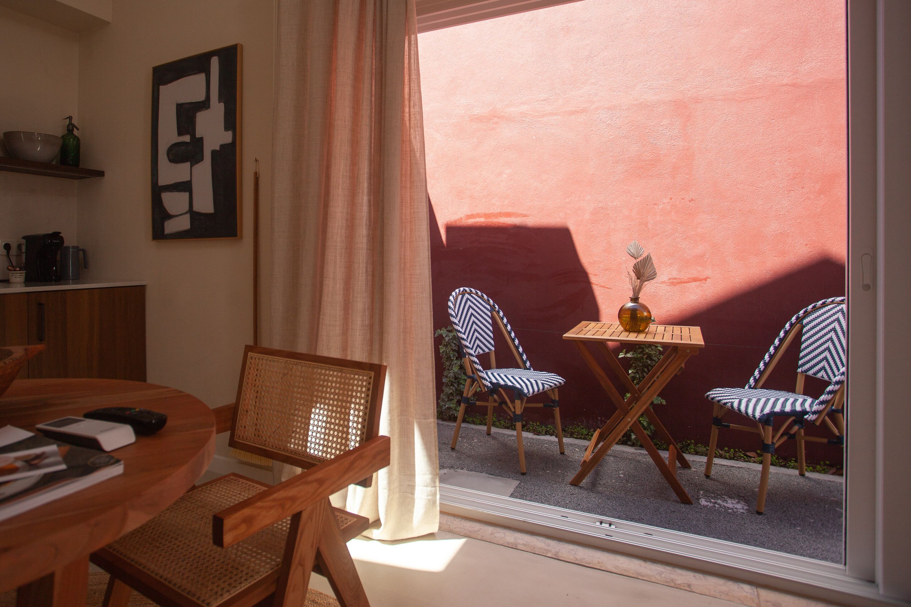 Una habitación con una mesa de comedor, sillas, una mesita con un florero y una ventana que ofrece vistas a un muro rojo y vegetación exterior.