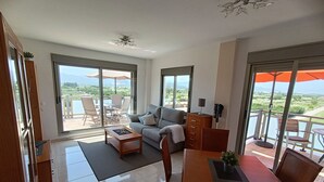 A spacious living area with a sofa, coffee table, and dining table, large sliding glass doors leading to a balcony with outdoor seating and a view of the landscape.