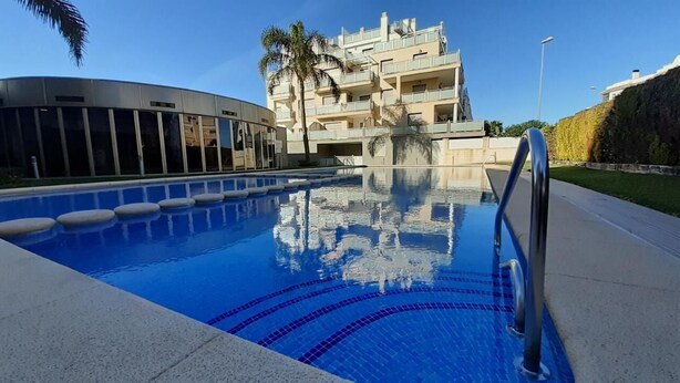 A swimming pool with clear blue water, a modern building with balconies, and a palm tree in the background.