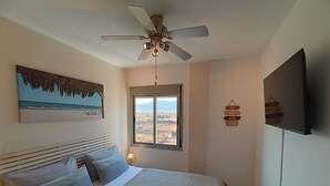 A bedroom with a bed, a ceiling fan, a window with a view, a painting, and a wall-mounted TV.