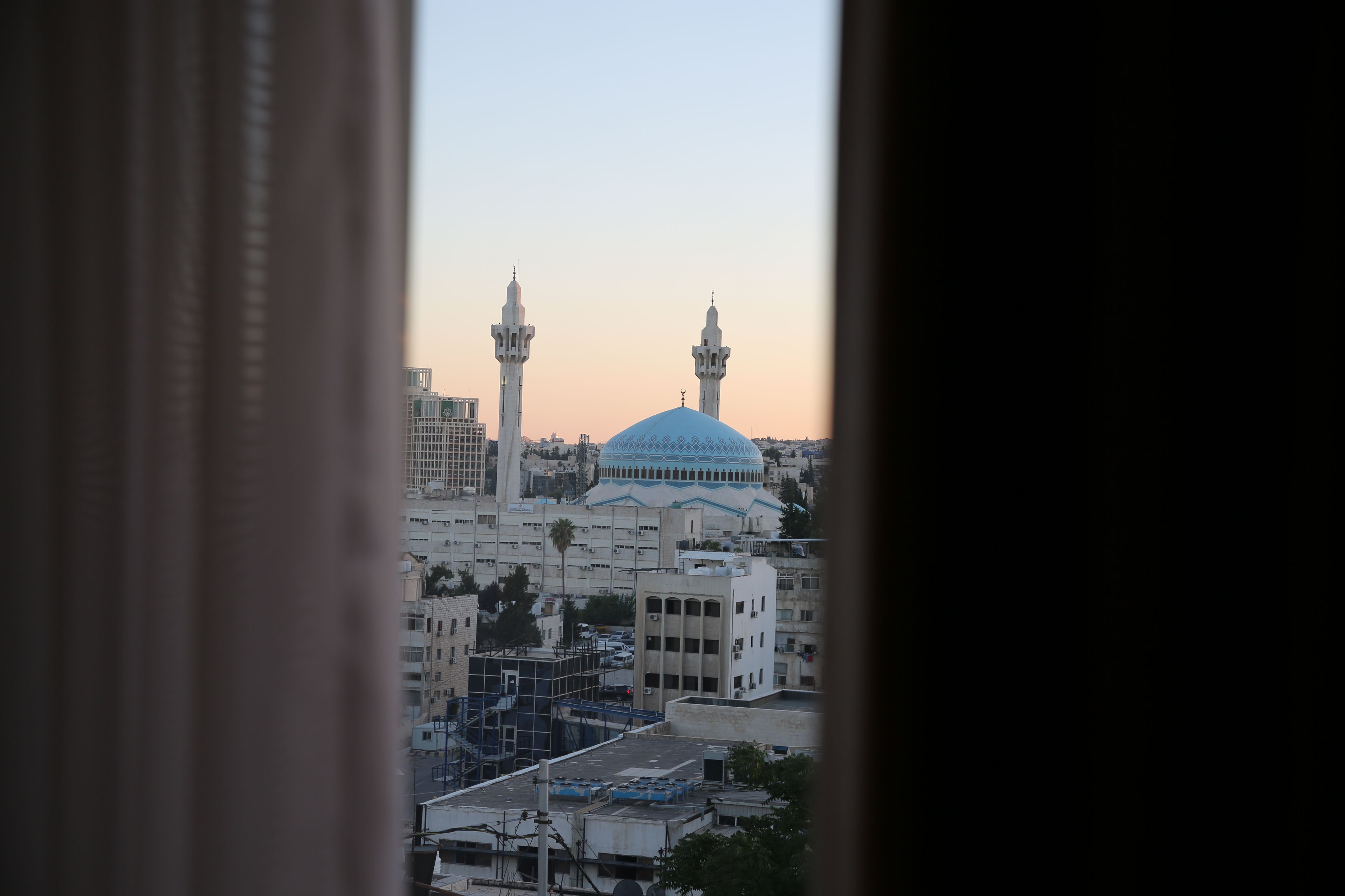 A view of a mosque with two minarets and a large dome, seen through a window with curtains.