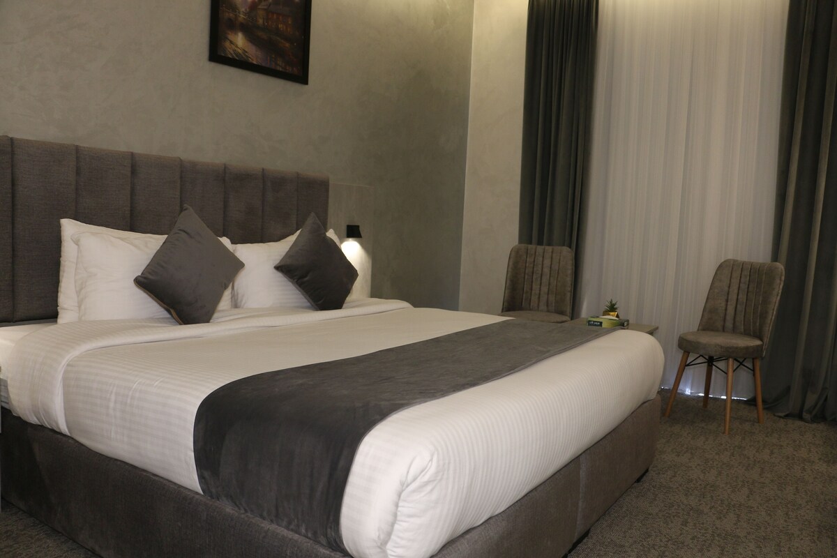 Una cama bien hecha con cabecera gris, una mesita de noche con lámpara y una silla en una habitación de hotel.