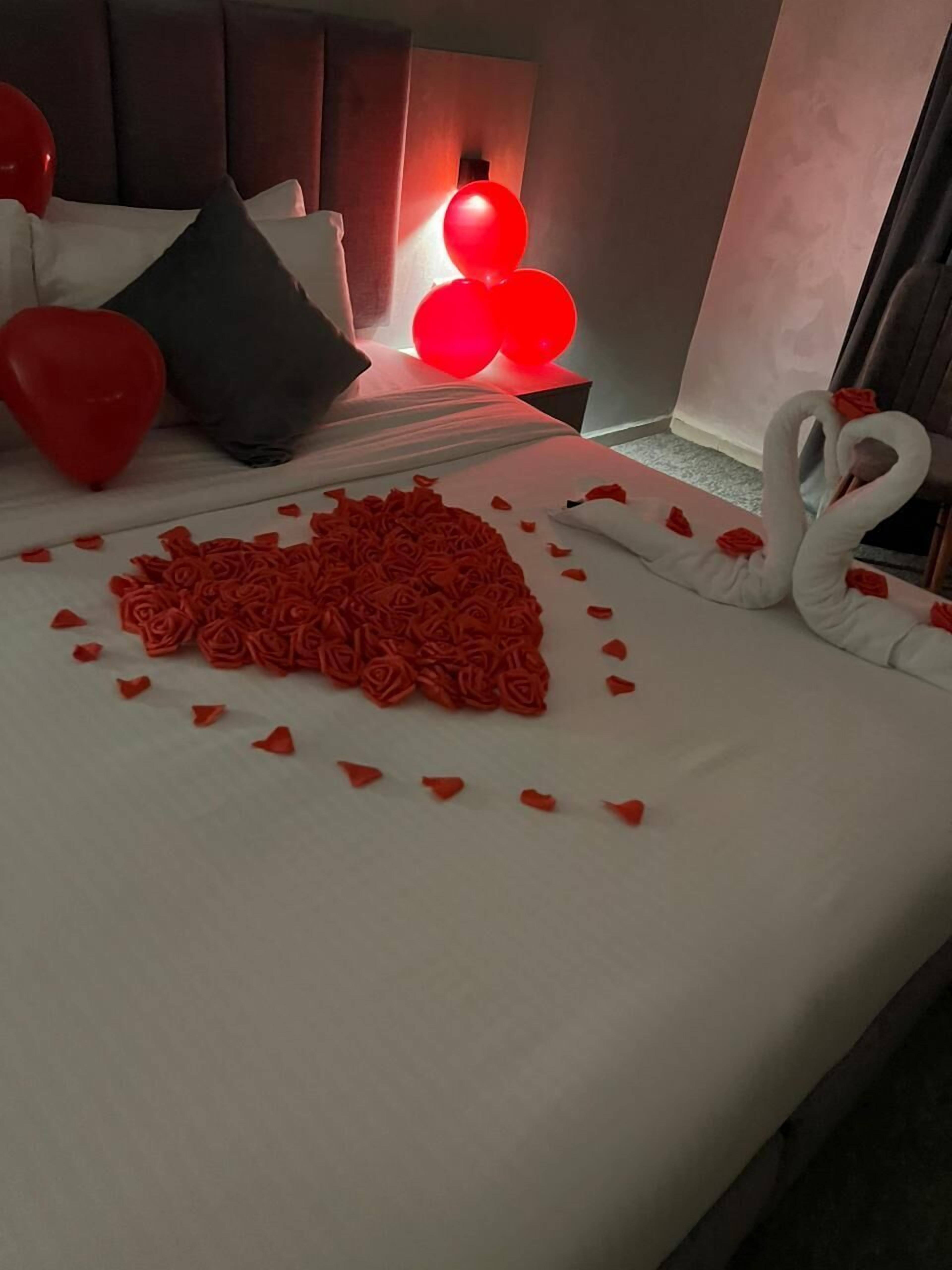 A bed with a heart-shaped arrangement of rose petals and swan-shaped decorations, a pillow, and red balloons.