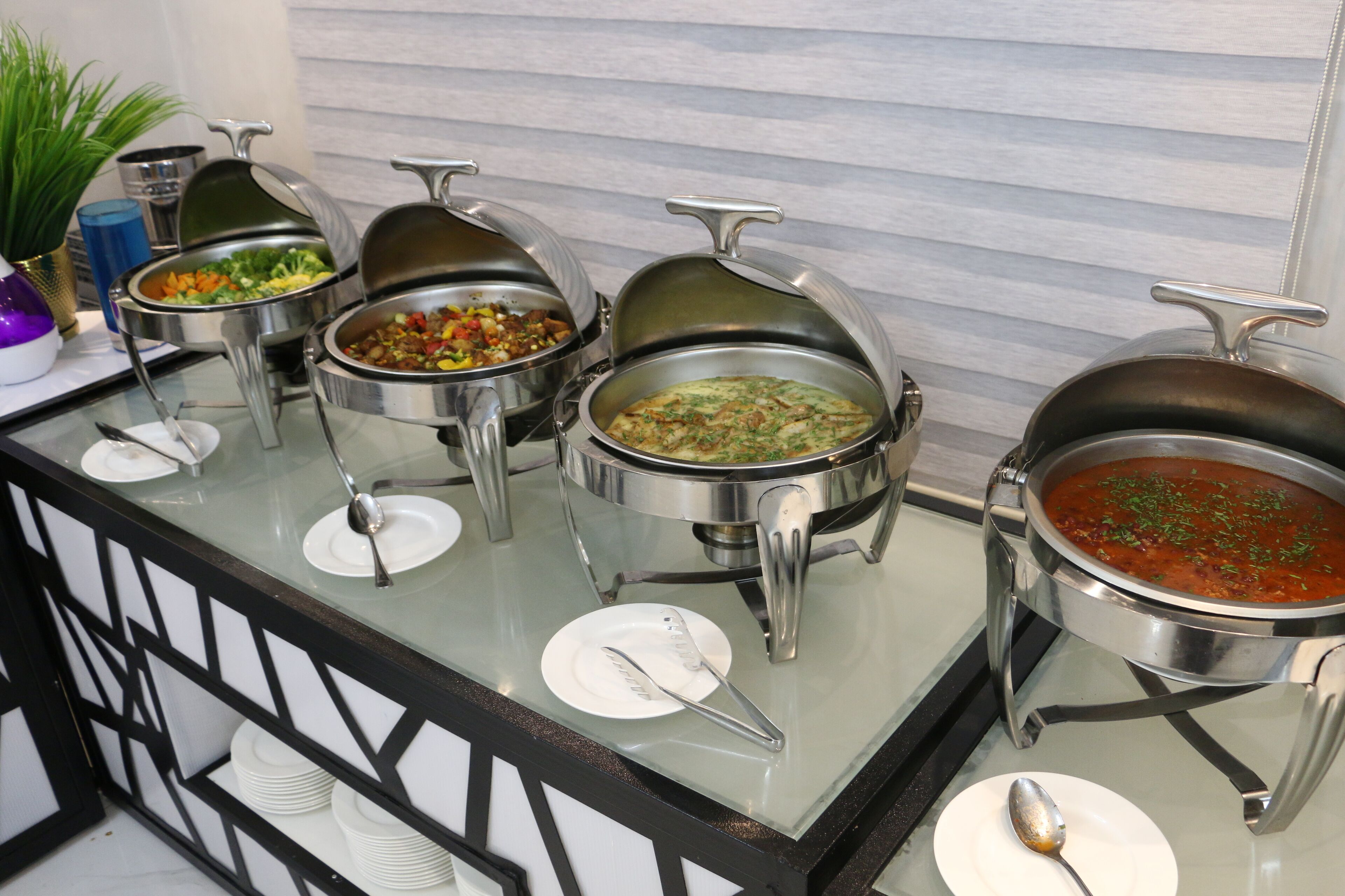 A buffet with various dishes in metal serving containers, a glass counter, and white plates with cutlery.