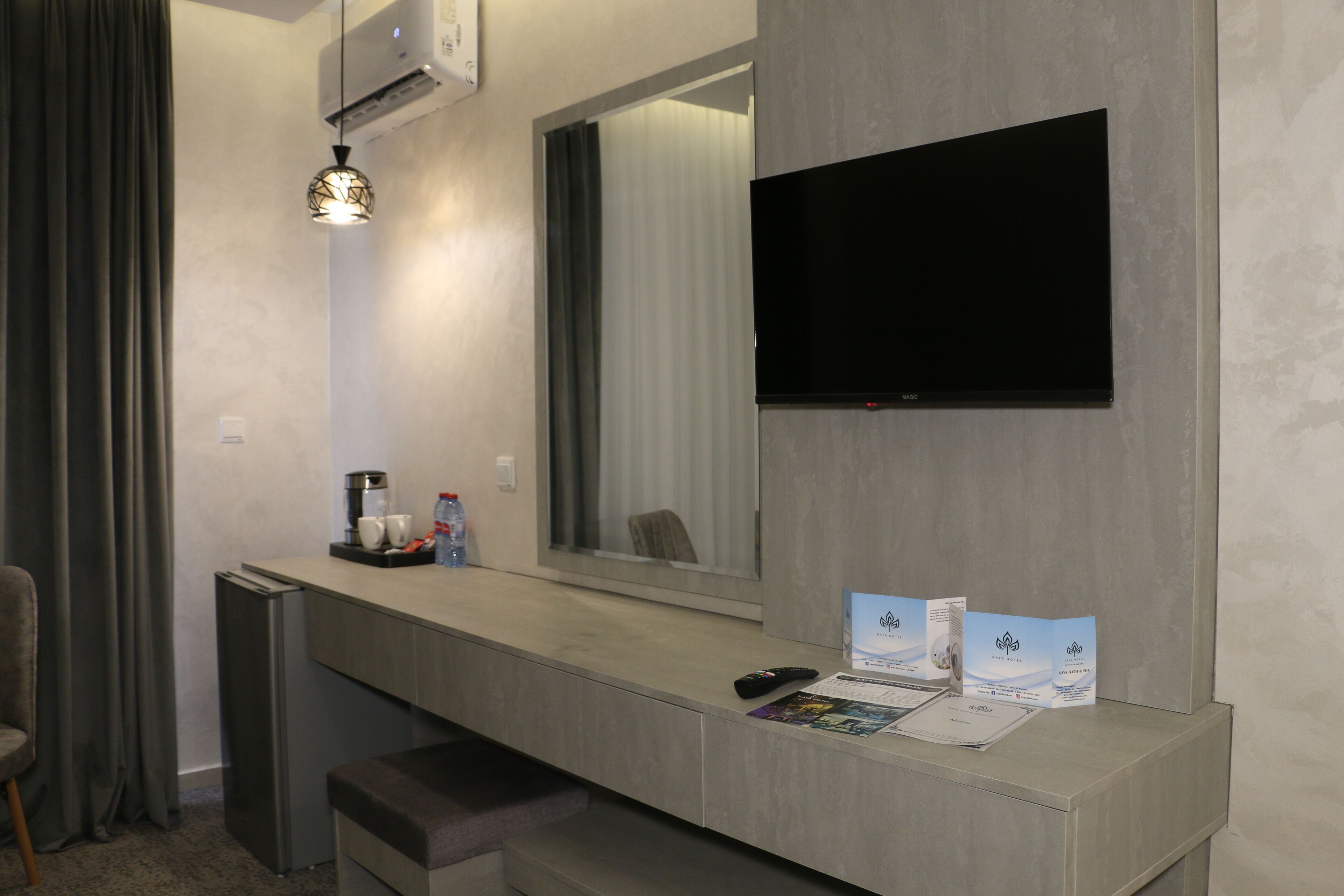 A hotel room with a modern kitchenette, a flat-screen TV mounted on the wall, a small seating area with a chair, and a counter with a coffee maker and brochures.