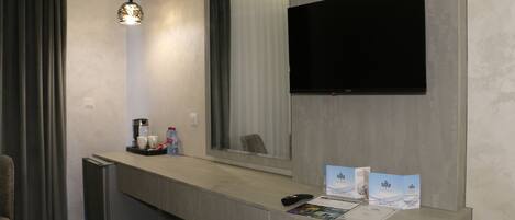 Habitación de hotel con kitchenette moderna, televisor de pantalla plana montado en la pared, un pequeño rincón de descanso con una silla y una encimera con cafetera y folletos.