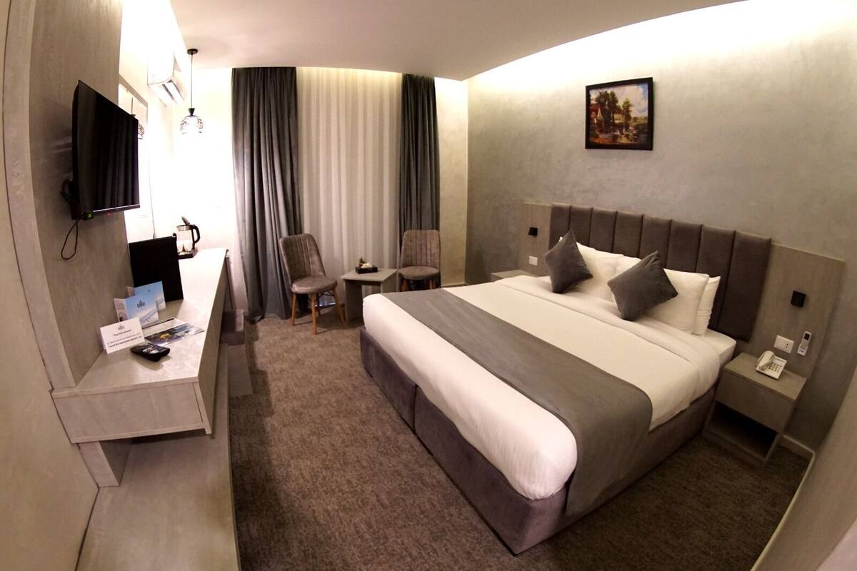 A hotel room with a large bed, a desk with a flat-screen TV, two chairs, a small table, and a painting on the wall.